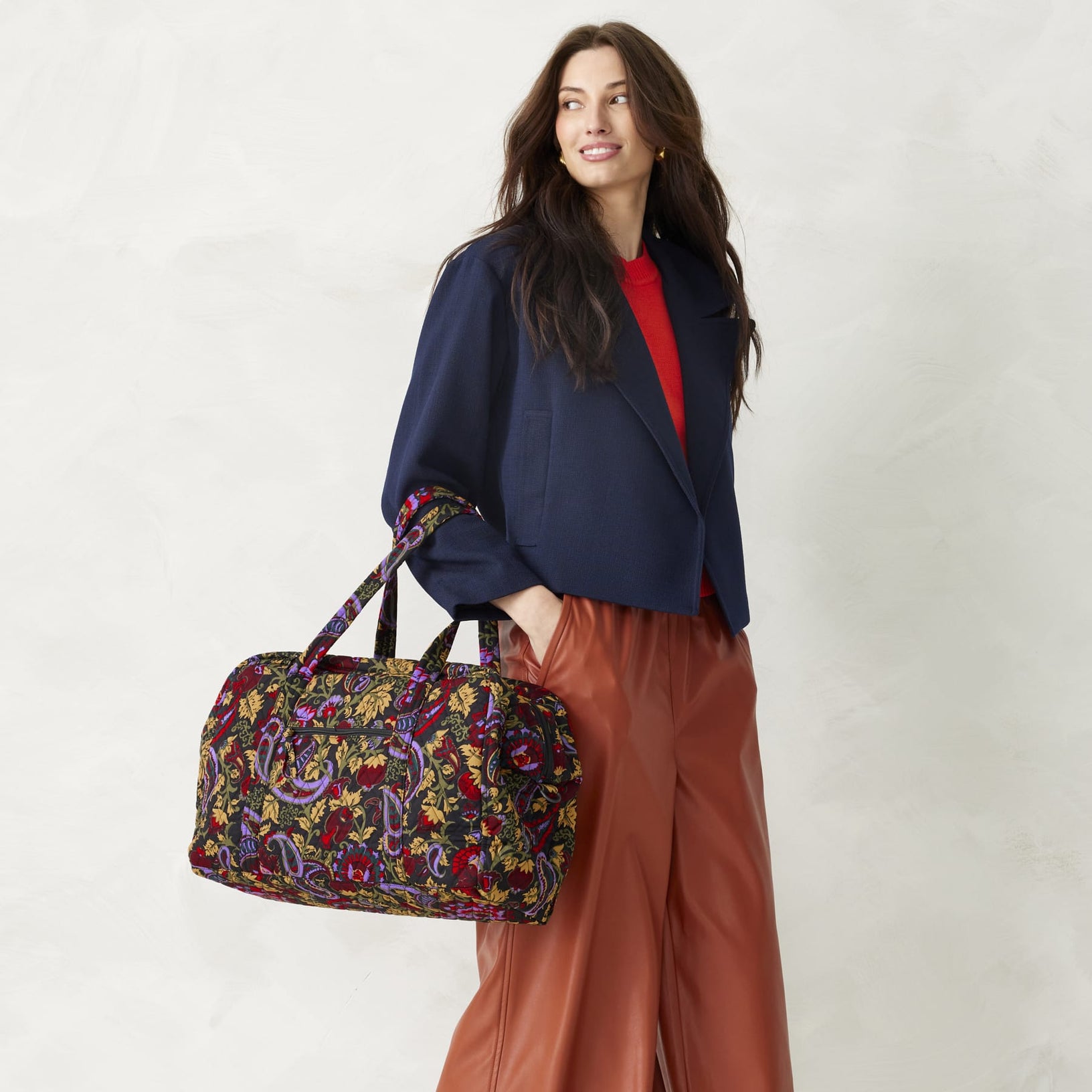 model wearing navy blue jacket carrying a Vera Bradley weekender bag with purple red and yellow paisley pattern