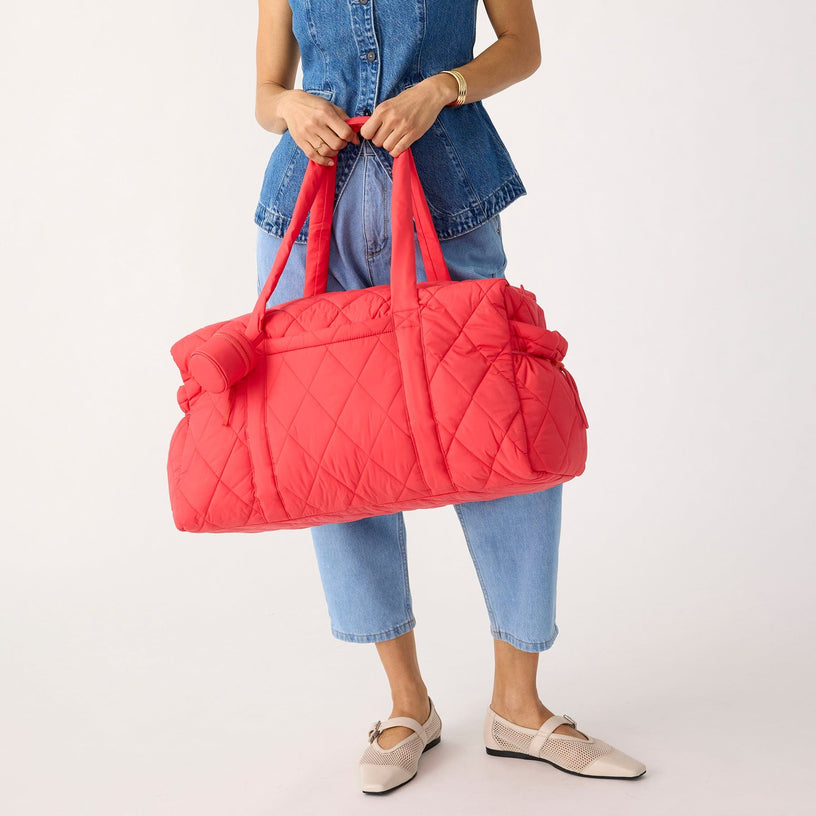 model wearing denim and holding large red quilted duffel bag with long handles